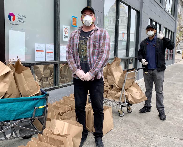 Two people in masks stand socially distanced next to multiple bags of groceries.