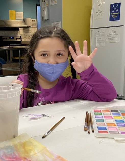 A child in a mask waves their hand sitting at a table with paints and paper.